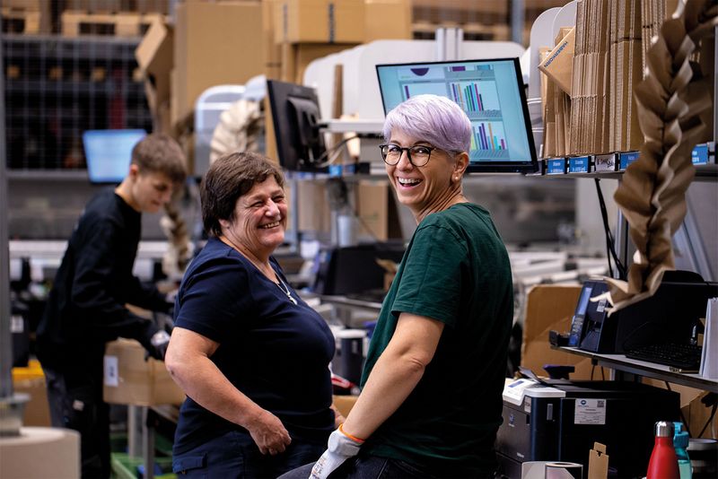 zwei Mitarbeiterinnen von Haberkorn in der Logistik lachen in die Kamera; im Hintergrund jüngerer Mitarbeiter bei der Arbeit