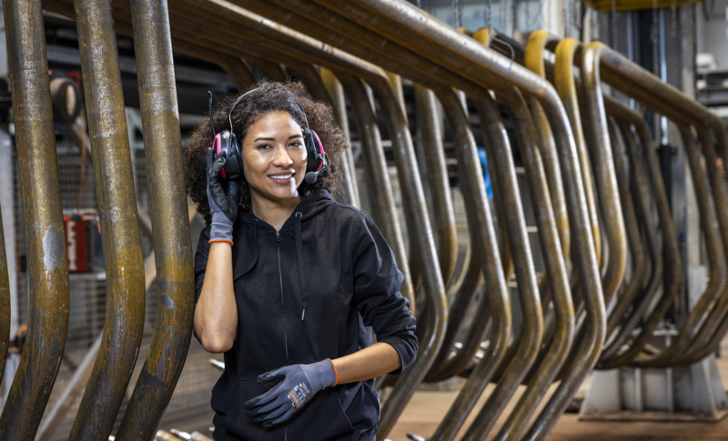 Frau in Arbeitskleidung steht vor Stahlteilen die von der Decke hängen und greift sich mit der rechten Hand an den Gehörschutzhörer den sie trägt.