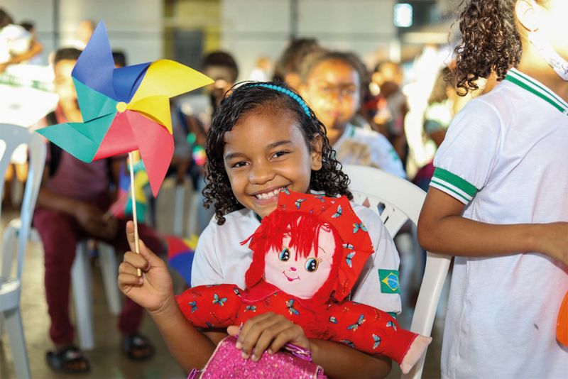 kleines Mädchen mit buntem Windrad und Puppe in der Hand lächelt aus einer Menschenmenge im Hintergrund